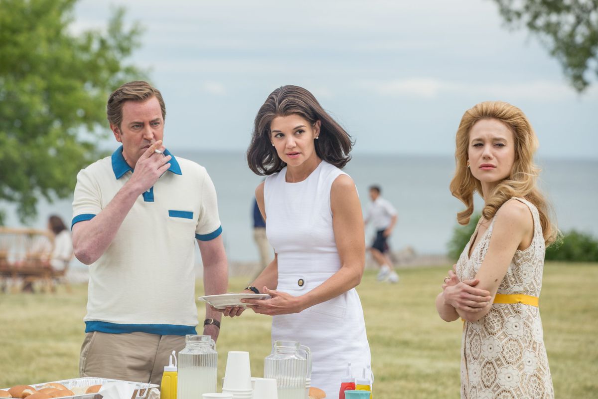 Ted Kennedy (Matthew Perry) smoking next to two women standing near a food table. They’re all looking at something off-screen