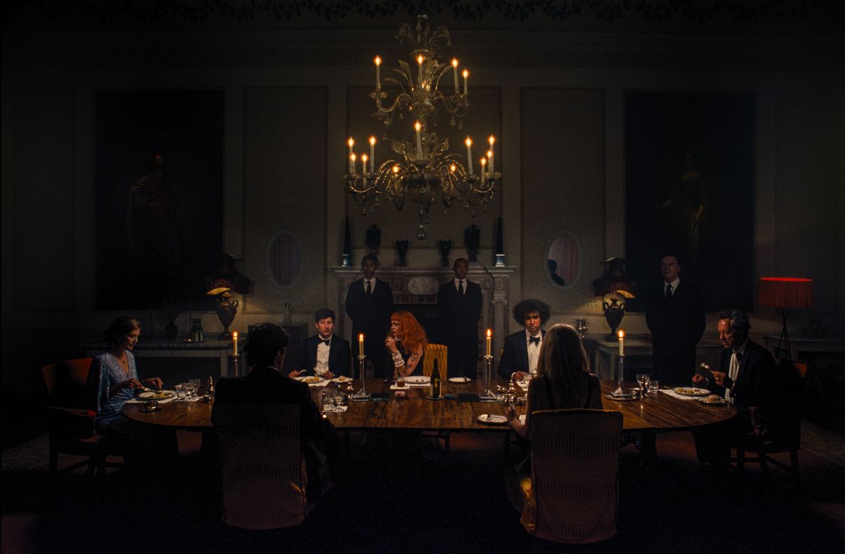 Oliver (Barry Keoghan) sits at a long dining-room table in a very dark room with Felix (Jacob Jacob Elordi) and other members of his family, all in formalwear, in the movie Saltburn
