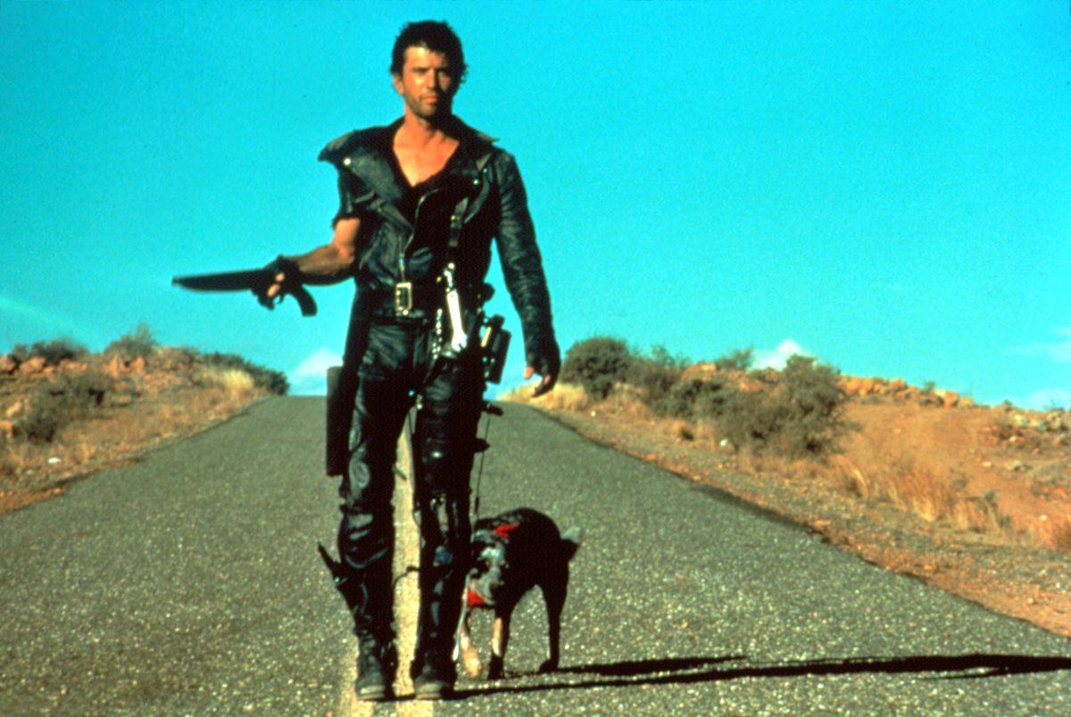 Mel Gibson as Mad Max, wearing all black leather and holding a short shotgun, walks with a dog on a road in Mad Max 2: The Road Warrior.