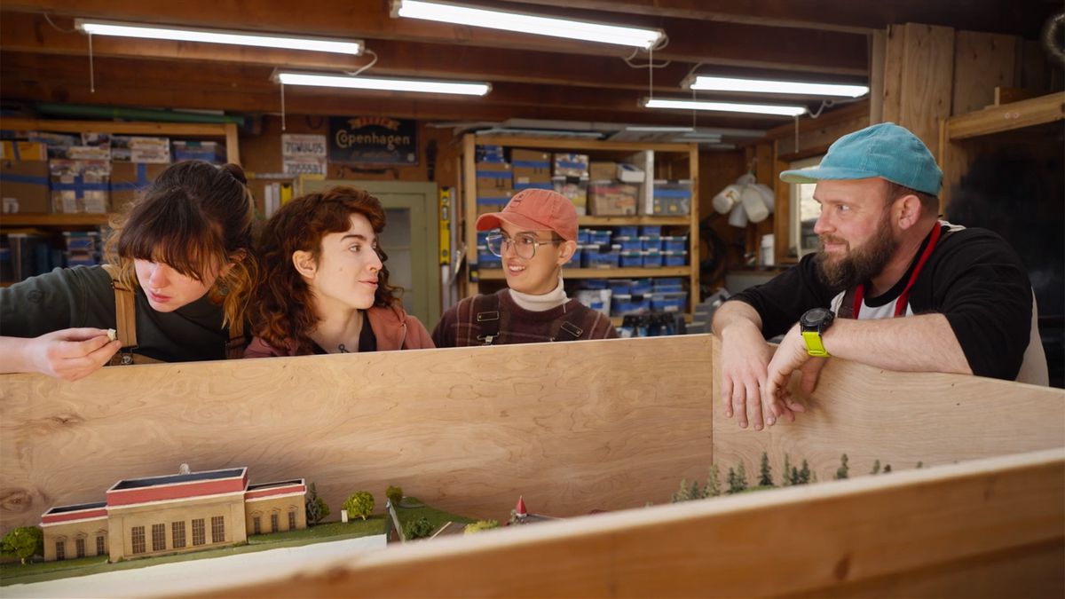 Rick Perry in a blue ball cap stands next to three of his teammates inside a rough hewn shop with exposed timbers. Bins of miniatures sit on shelves in the background.