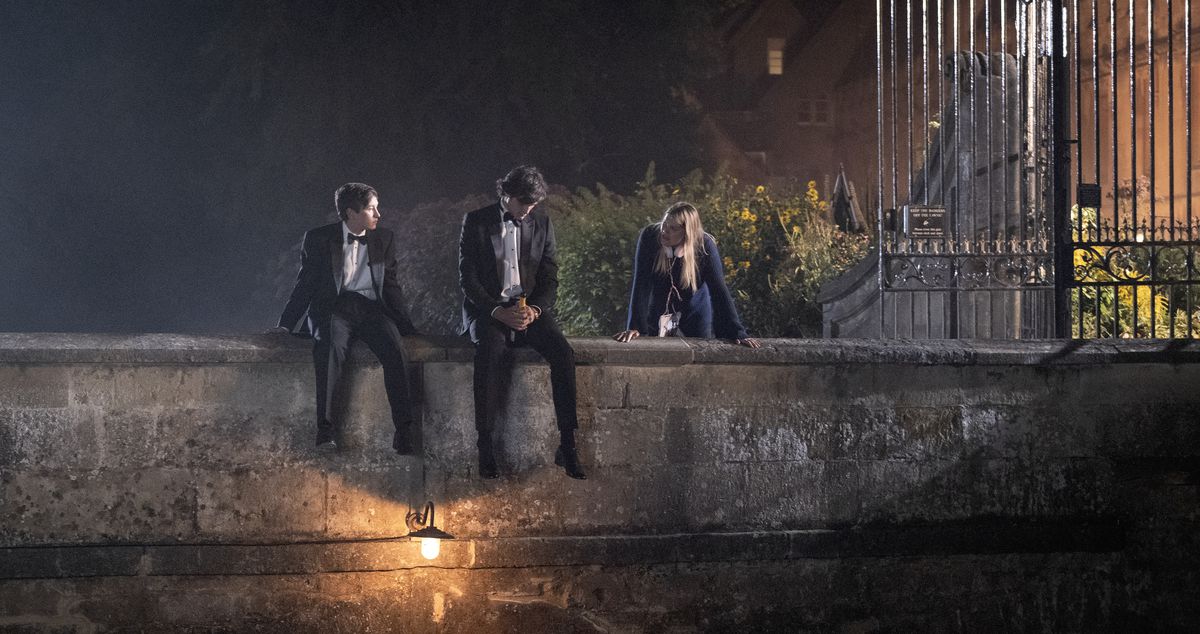 Oliver (Barry Keoghan) and Felix Catton (Jacob Elordi), in tuxes, sit together on a small stone bridge over a pond with Venetia (Alison Oliver) standing nearby in Saltburn
