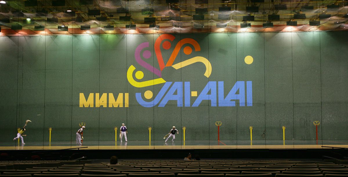 Players throw balls using hook shaped baskets on their arms in a large Jai alai court, with the words “Maimi Jai Alai” artistically depicted on the background wall.