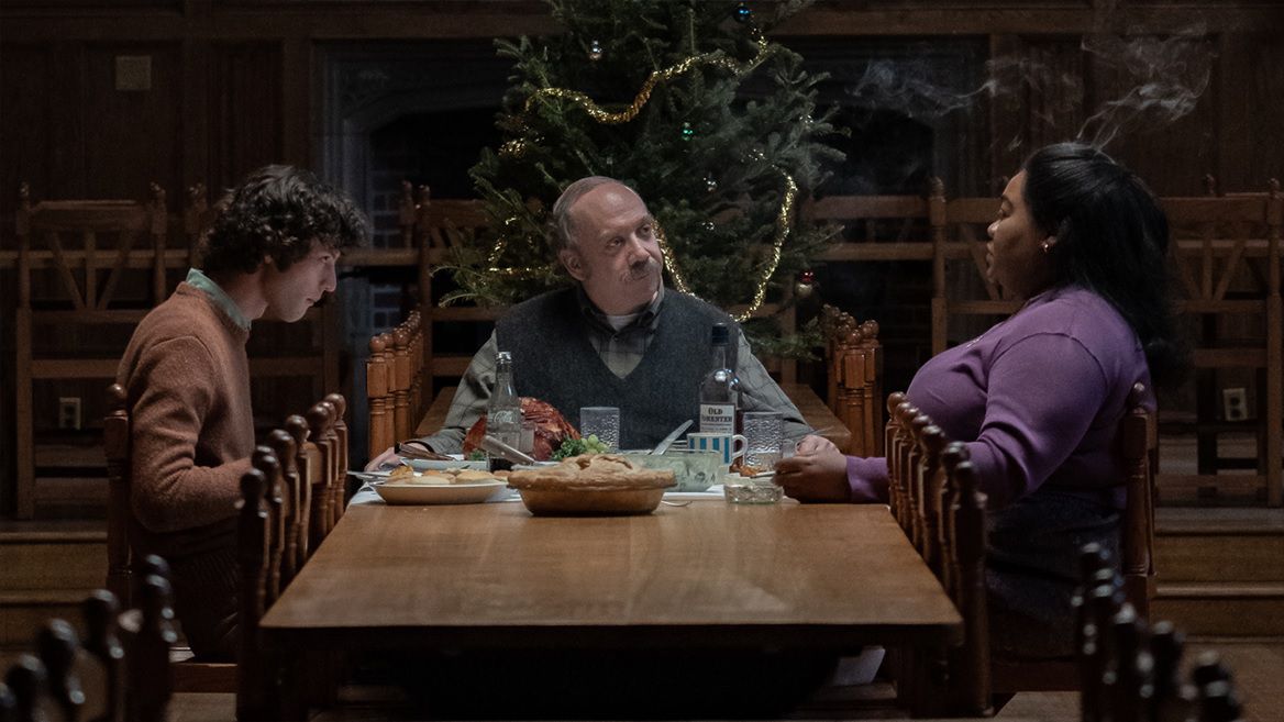 Dominic Sessa, Paul Giamatti, and Da’Vine Joy Randolph gather around a table with a Christmas tree in the background in The Holdovers.