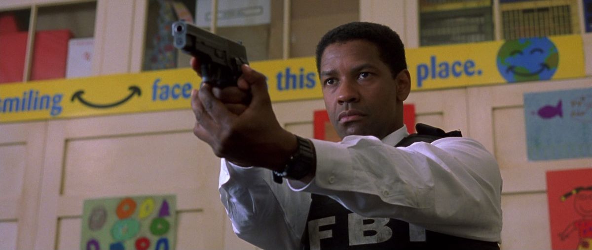 Denzel Washington wears a bulletproof vest that says “FBI” and aims a gun while standing in what looks like a school gym in The Siege.