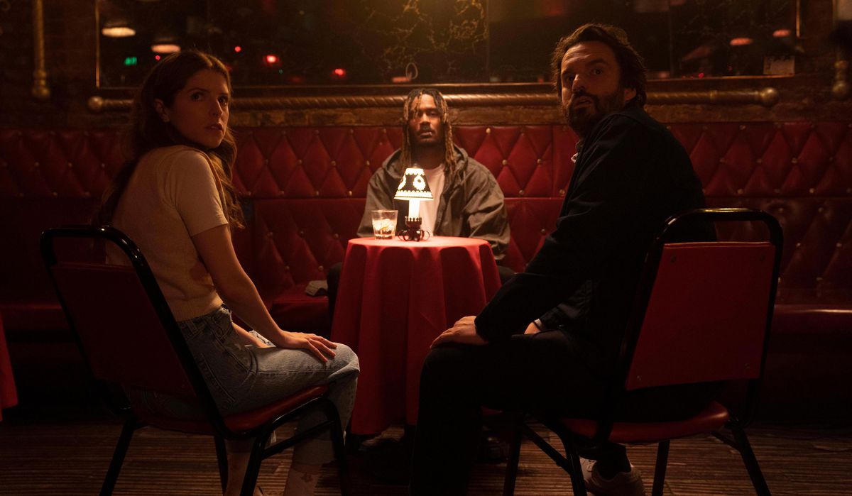 Maddie (Anna Kendrick) and Tommy (Jake Johnson) look nervously over their shoulders as they sit in a dark bar upholstered in red, opposite a tiny table where Charlie (GaTa) sits in front of a tiny light in Johnson’s movie Self Reliance