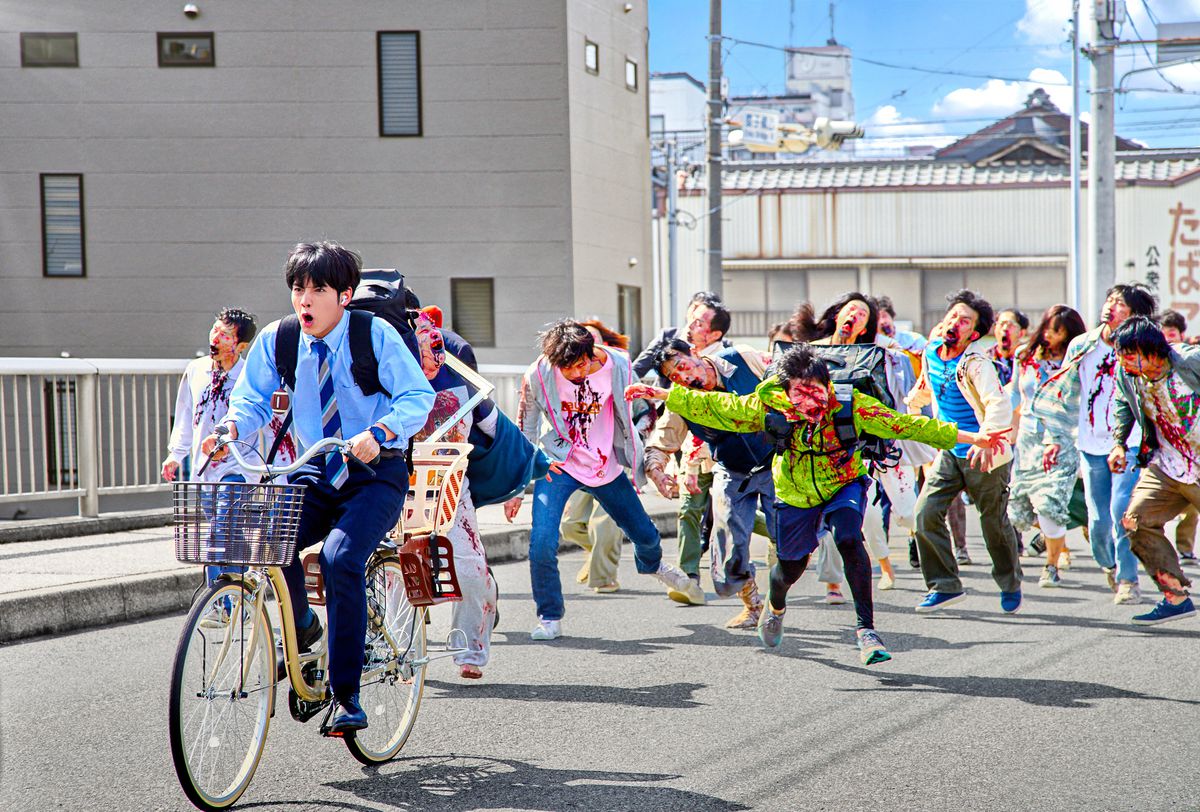 Eiji Akaso as Akira riding a bicycle away from a horde of zombies in Zom 100: Bucket List of the Dead.