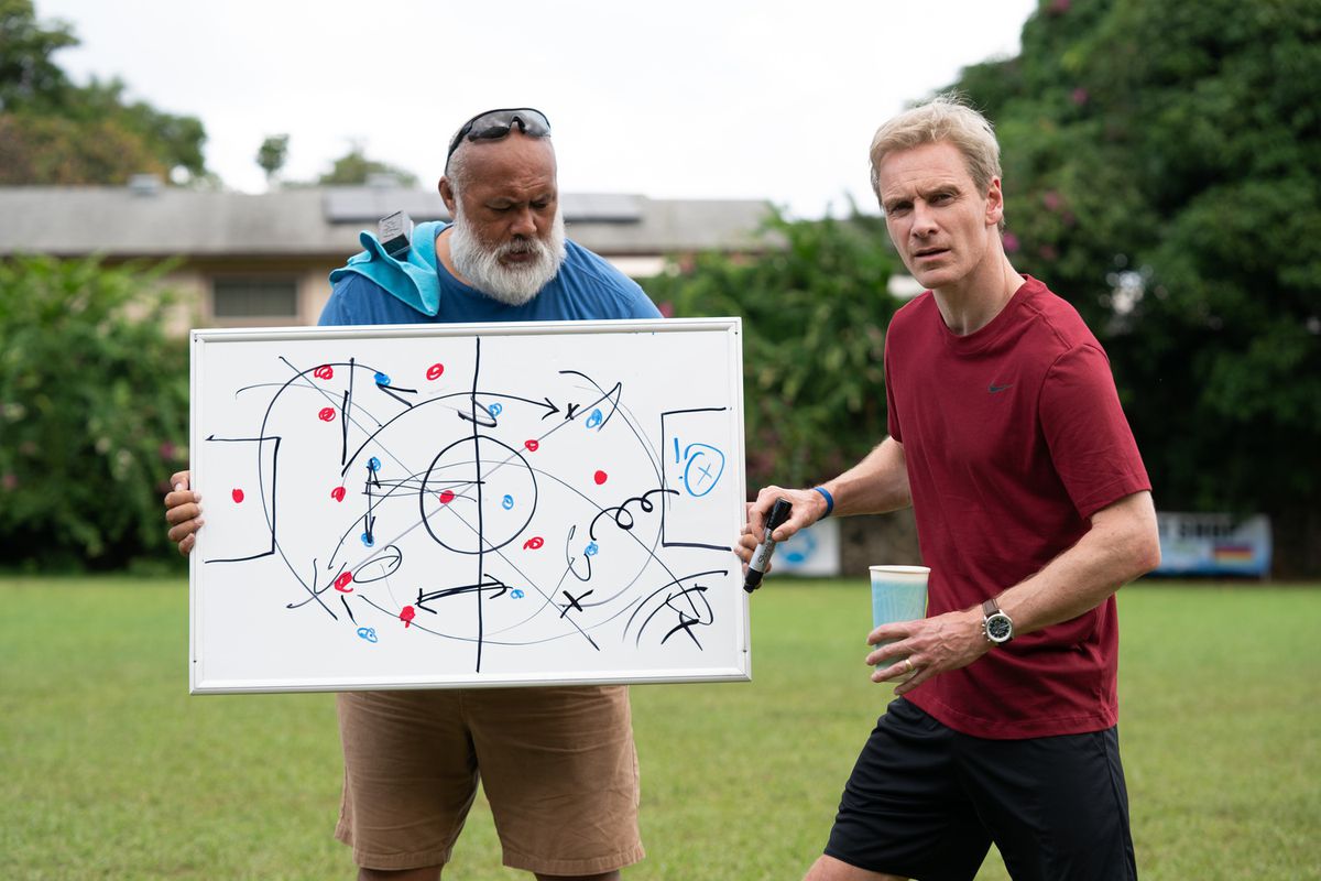 Ace (David Fane) holding a whiteboard while coach Thomas Rongen (Michael Fassbender) lectures his team off-screen in Next Goal Wins.