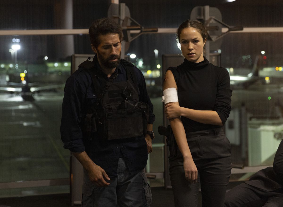 Scott Adkins stands next to a wounded Hannah Arterton, with a bandage on her arm, in the airport in One More Shot.