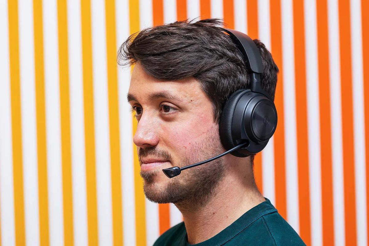 The SteelSeries Arctis Nova Pro Wireless being worn by the author, Cameron Faulkner. He is standing in front of a wall that has a gradient pattern going from warm yellow to orange.