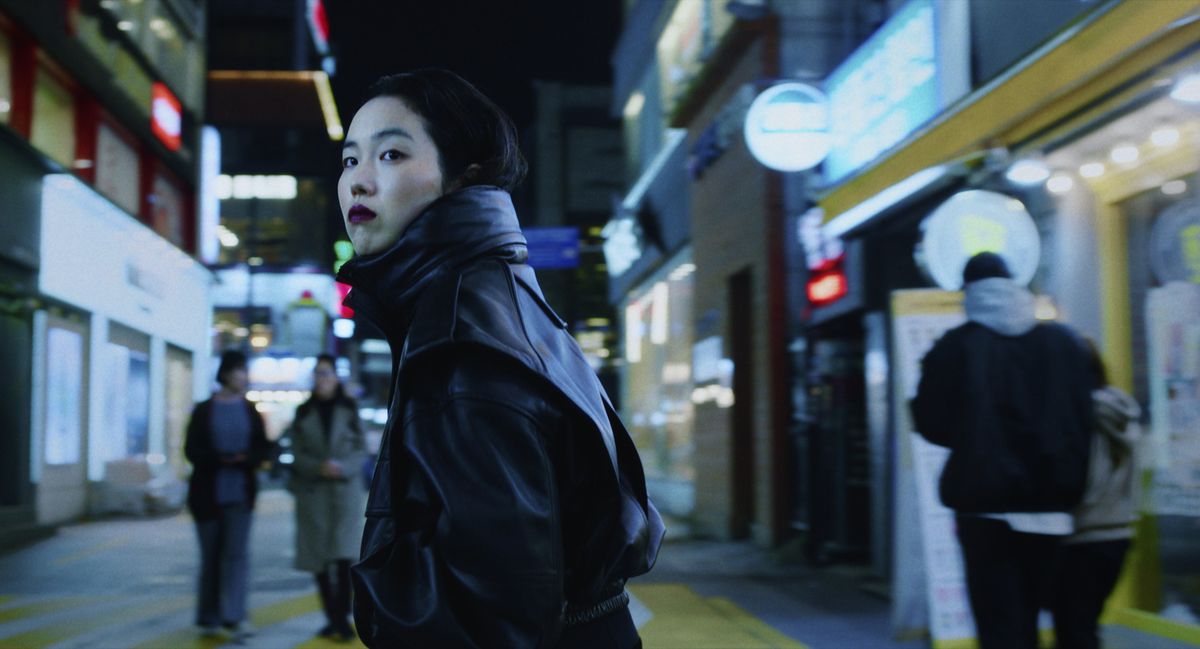 Jin-Min Park as Freddie, wearing dark red lipstick in a black leather coat standing in the middle of a Korean city street at night in Return to Seoul.