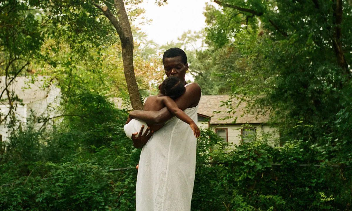 A woman (Sheila Atim) in a white dress cradling a child in their arms in front of a thicket of trees and bushes with a house in the background in All Dirt Roads Taste of Salt.