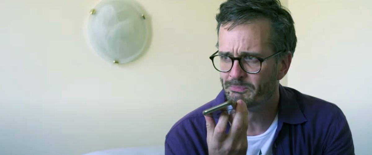 Journalist David Farrier sits on a bed in a white-walled room, holding a cell phone under his chin, and cries in a scene from his documentary Mister Organ