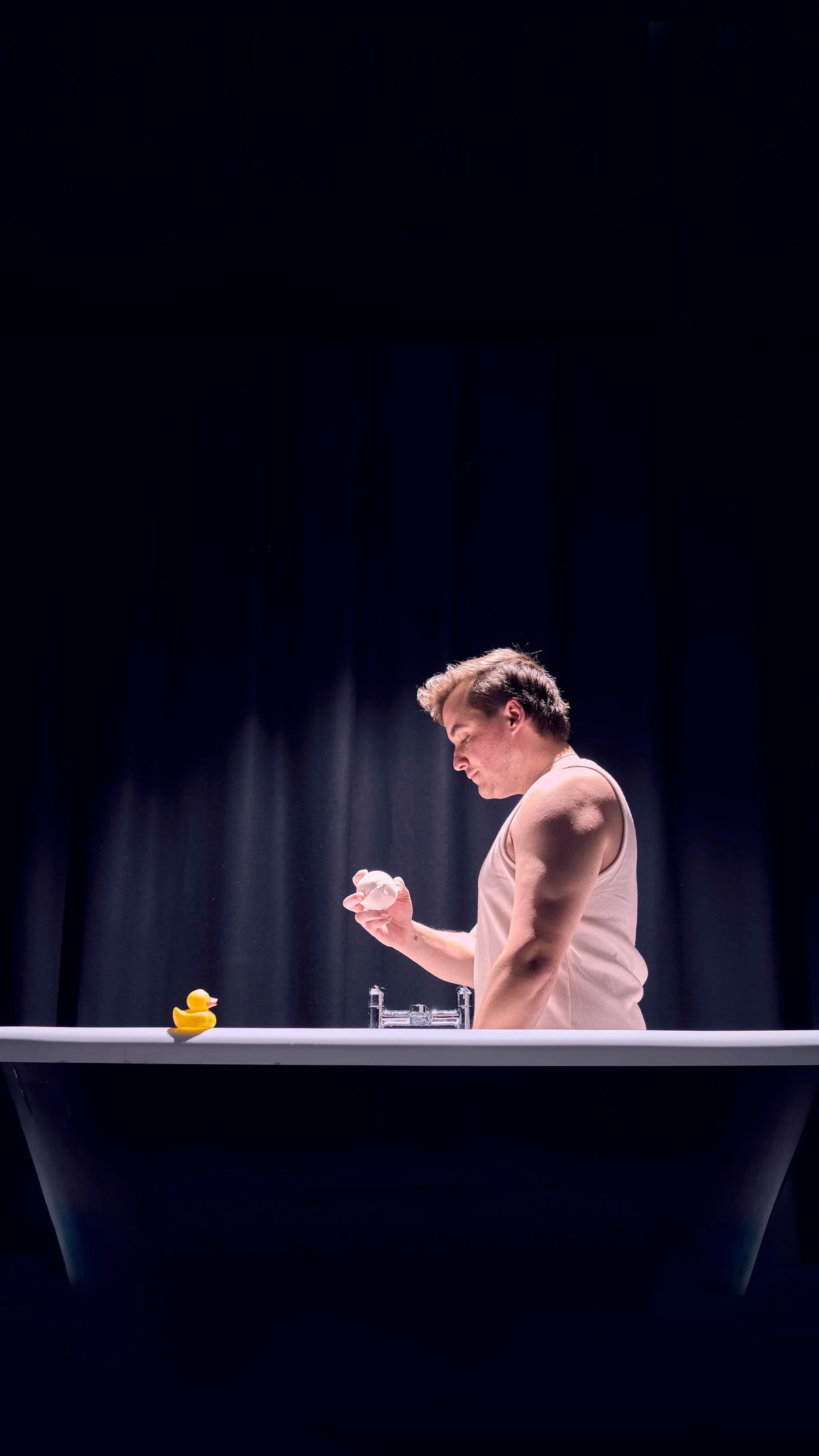 A man stands in a dramatically lit bathtumb. He wears a white tanktop and holds a white bathbomb, looking at it intensely. 
