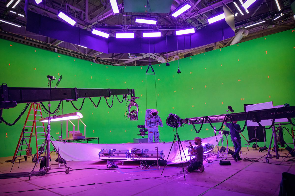 A figure in a spacesuit is dwarfed by cranes, cameras, and equipment in a brightly light green-and-purple high-ceilinged space on the set of Netflix’s Spaceman