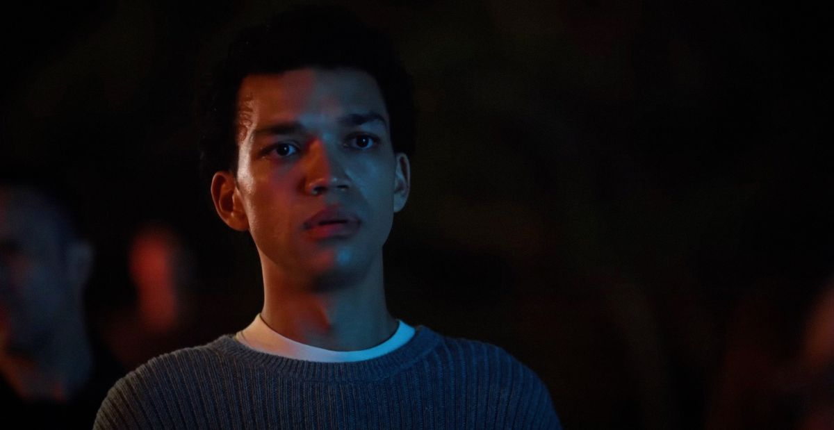 Owen (Justice Smith) stands in darkness with his face symbolically divided in half, with orange light on one side and a sickly blue-ish light on the other in Jane Schoenbrun’s movie I Saw the TV Glow 
