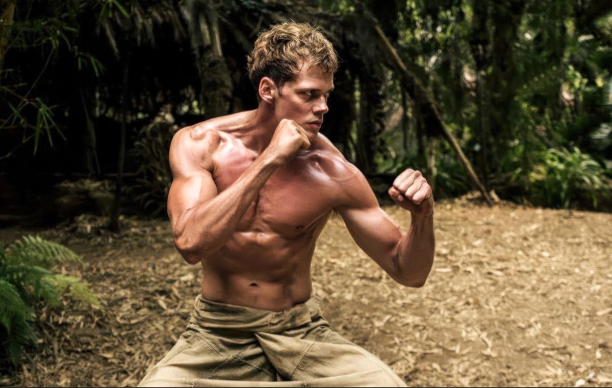 Boy (Bill Skarsgård) stands in a jungle clearing, shirtless and oiled-up and muscular, in a side-on, fists-up, ready-to-fight position straight out of a fighting game in Boy Kills World