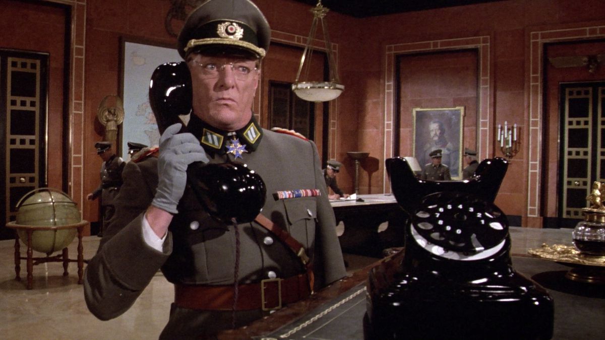 A man in a military uniform holding the receiver of a comically oversized rotary telephone to his ear and looking sternly.