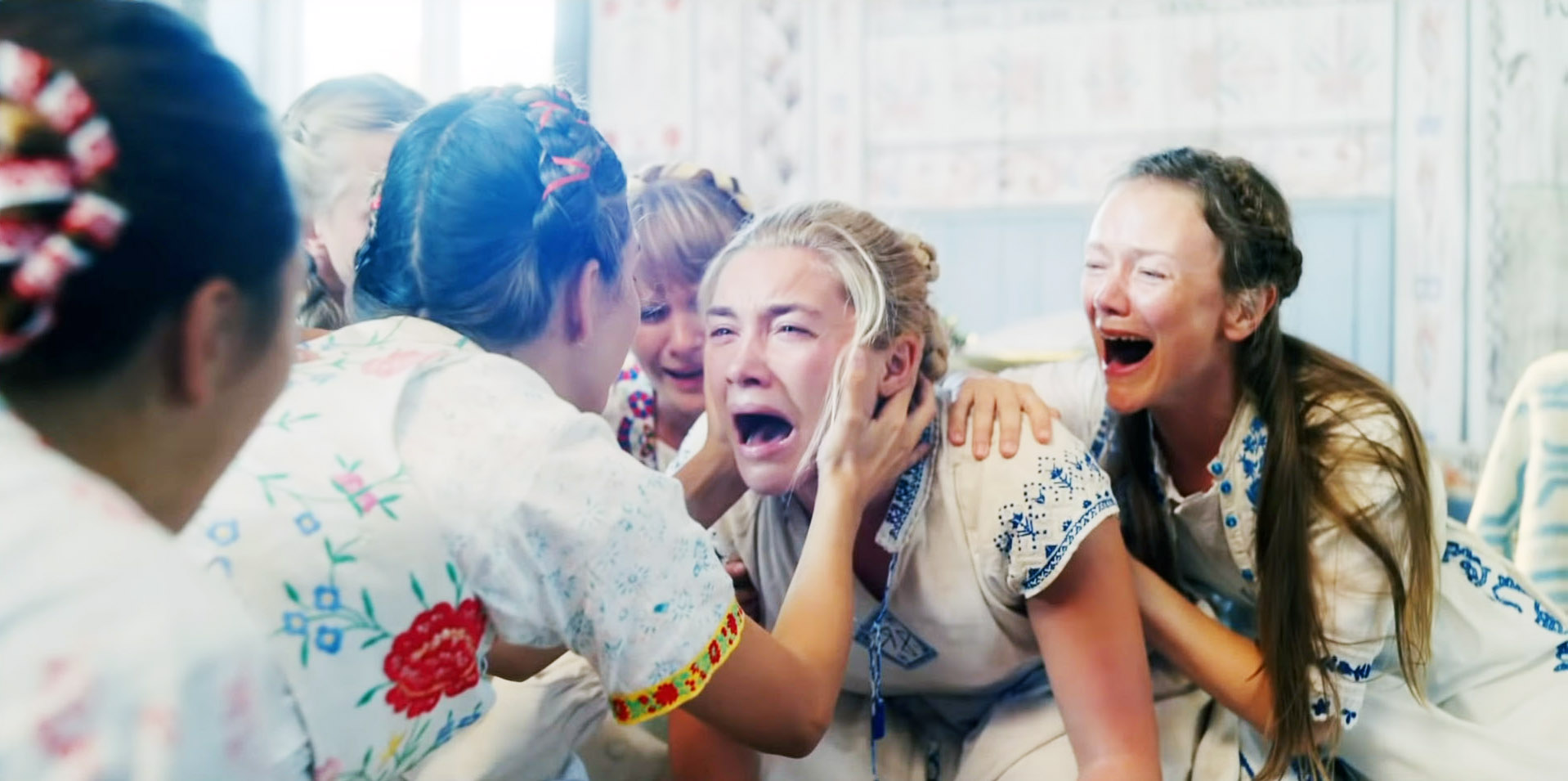 Dani (Florence Pugh), in a white dress with lace trim, kneels and wails amid a circle of similarly dressed women who are also wailing in Midsommar