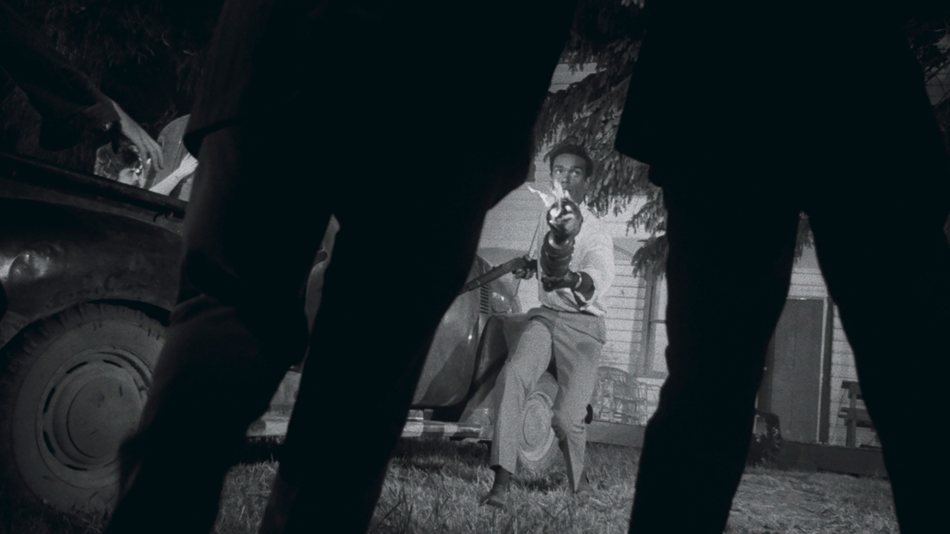 Ben (Duane Jones), a tall, thin Black man in a button-up white shirt, fires a gun at two silhouetted figures in the extreme foreground, blocking out most of the screen, in George A. Romero’s Night of the Living Dead