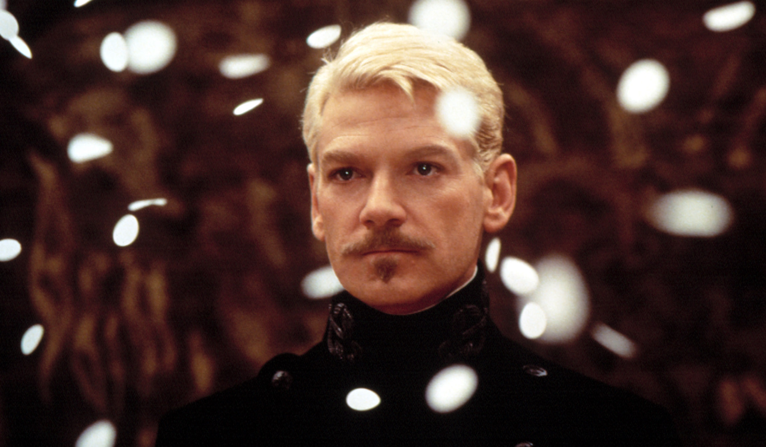 Kenneth Branagh as Hamlet, with a small pointed soul patch and bleached-blond hair, standing and frowning slightly as white circular confetti rains down on him in the 1996 Hamlet