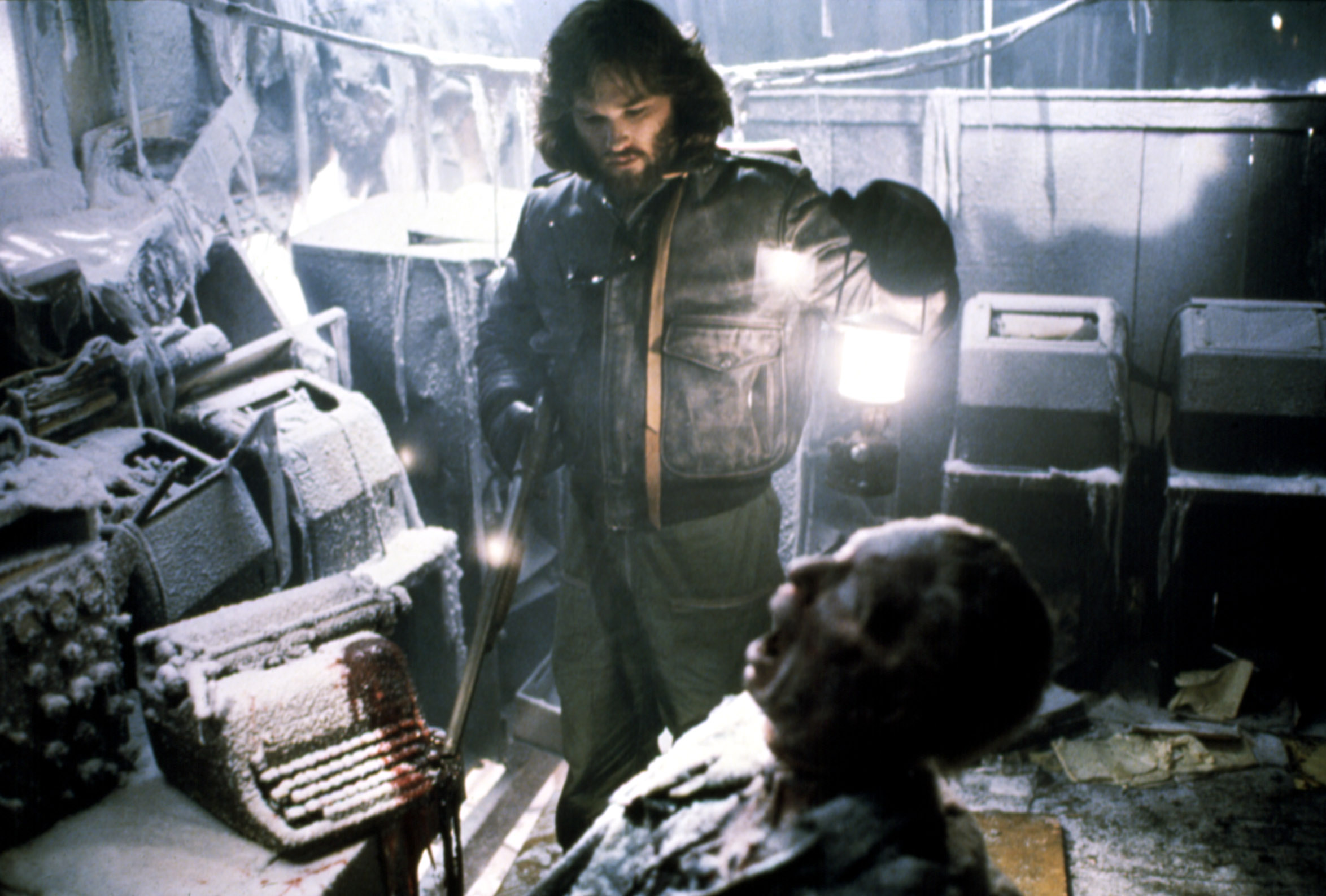 Macready (Kurt Russell), a man with shaggy hair and beard, wearing a full-body jumpsuit, holds up a lantern and examines a screaming frozen corpse in a blood-spattered, frozen room in the 1982 movie The Thing