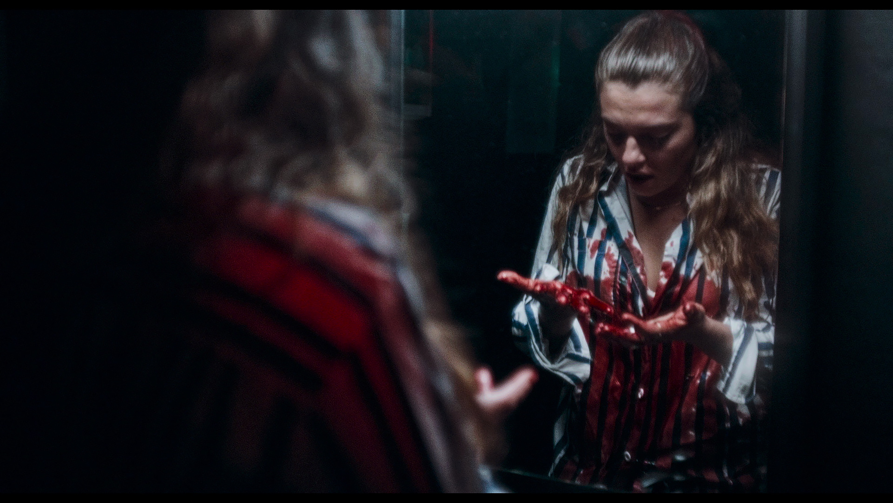 A young woman stands in a bathroom opposite a mirror, looking with horror at her blood-covered hands and shirt, in the French horror-thriller MadS