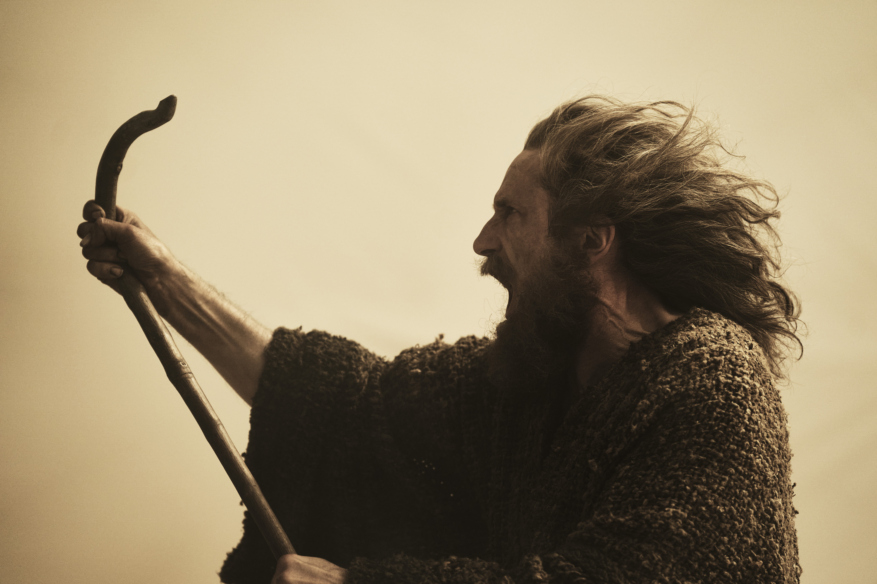 Daniel Weyman as The Stranger in The Lord of the Rings: The Rings of Power. He’s brandishing a staff-like branch into a strong wind and shouting.