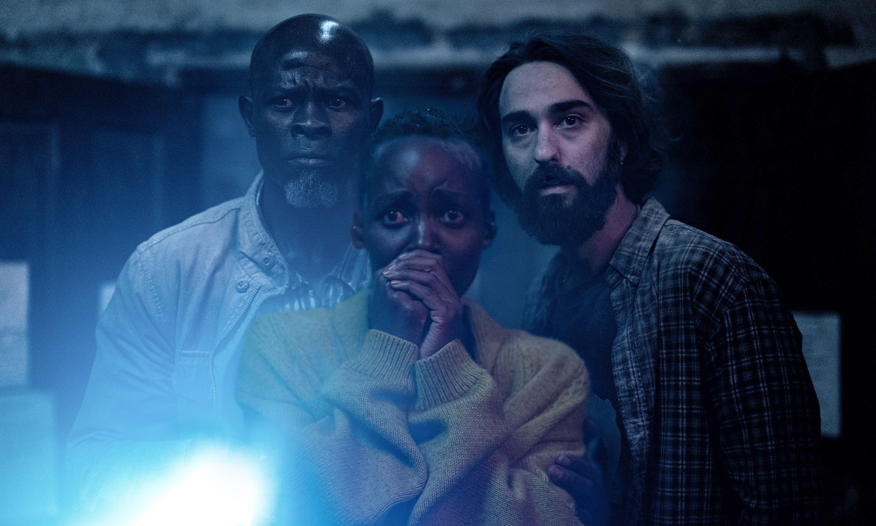 Three survivors of an alien invasion (Djimon Hounsou, Lupita Nyong’o, Alex Wolff) stand together in the dark, shining a flashlight toward the camera, in Michael Sarnoski’s A Quiet Place: Day One