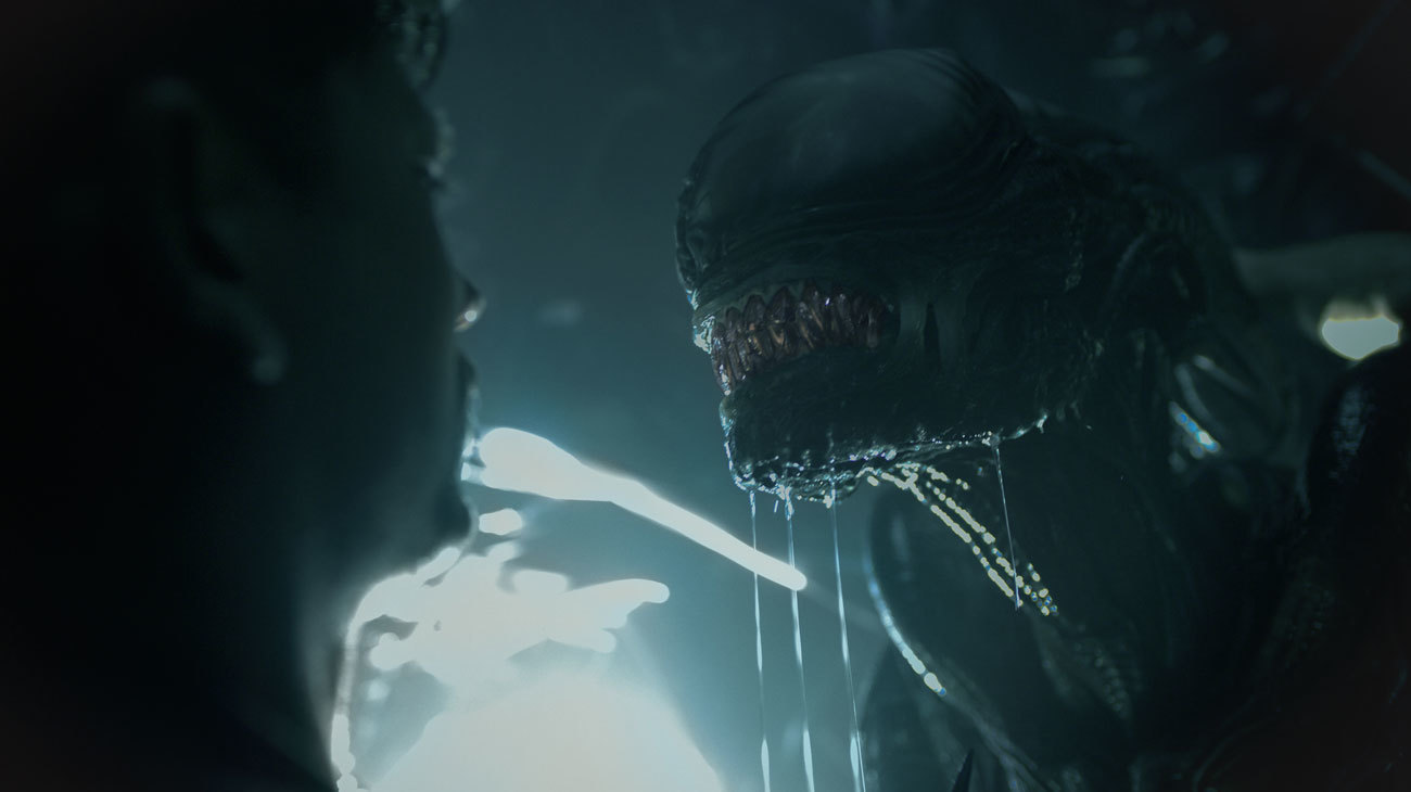 A close-up shot of a man staring at an eyeless alien creature with bared teeth and a drool-covered chin in Alien: Romulus.