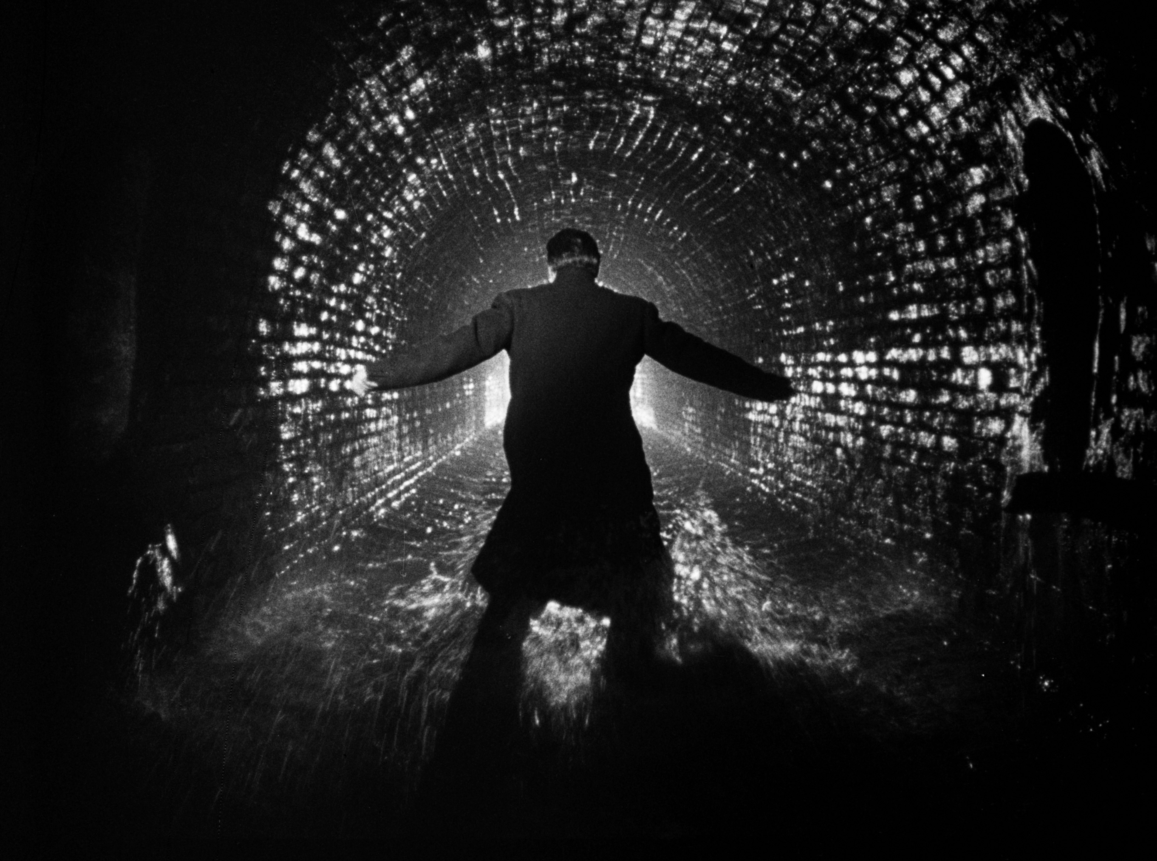 Orson Welles stands in a sewer, with his arms stretched out and facing away from the camera, in The Third Man