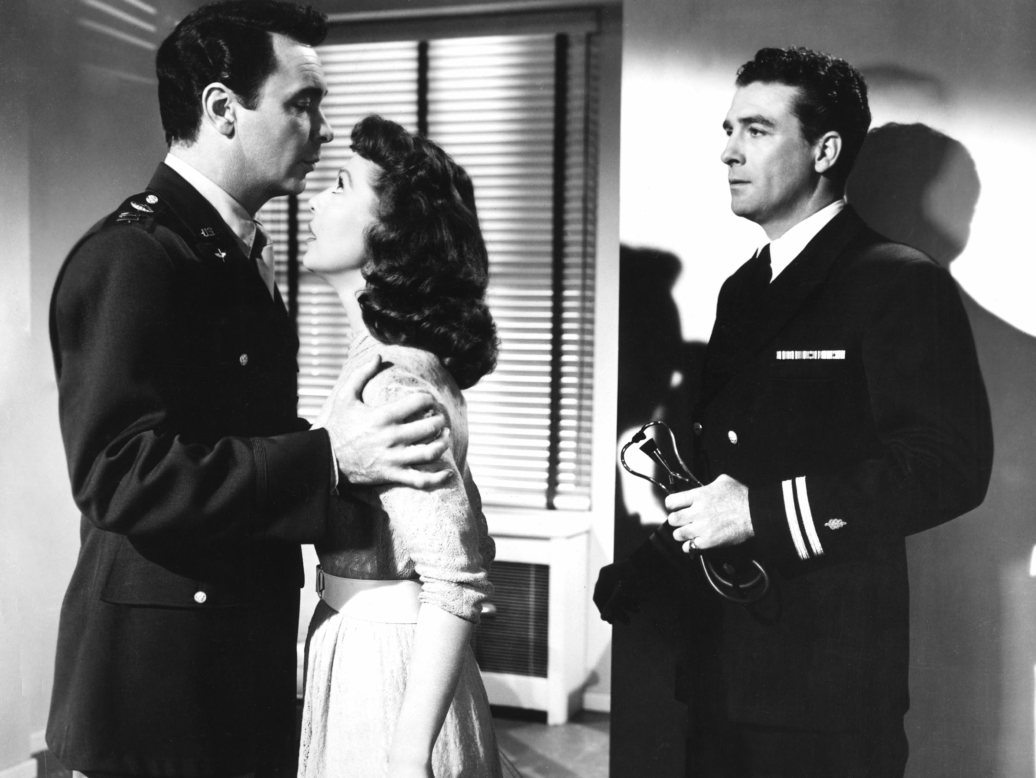 In Cause for Alarm!, Loretta Young stands between two men wearing military uniforms, as one has his arms on her shoulders. She looks deeply into his eyes