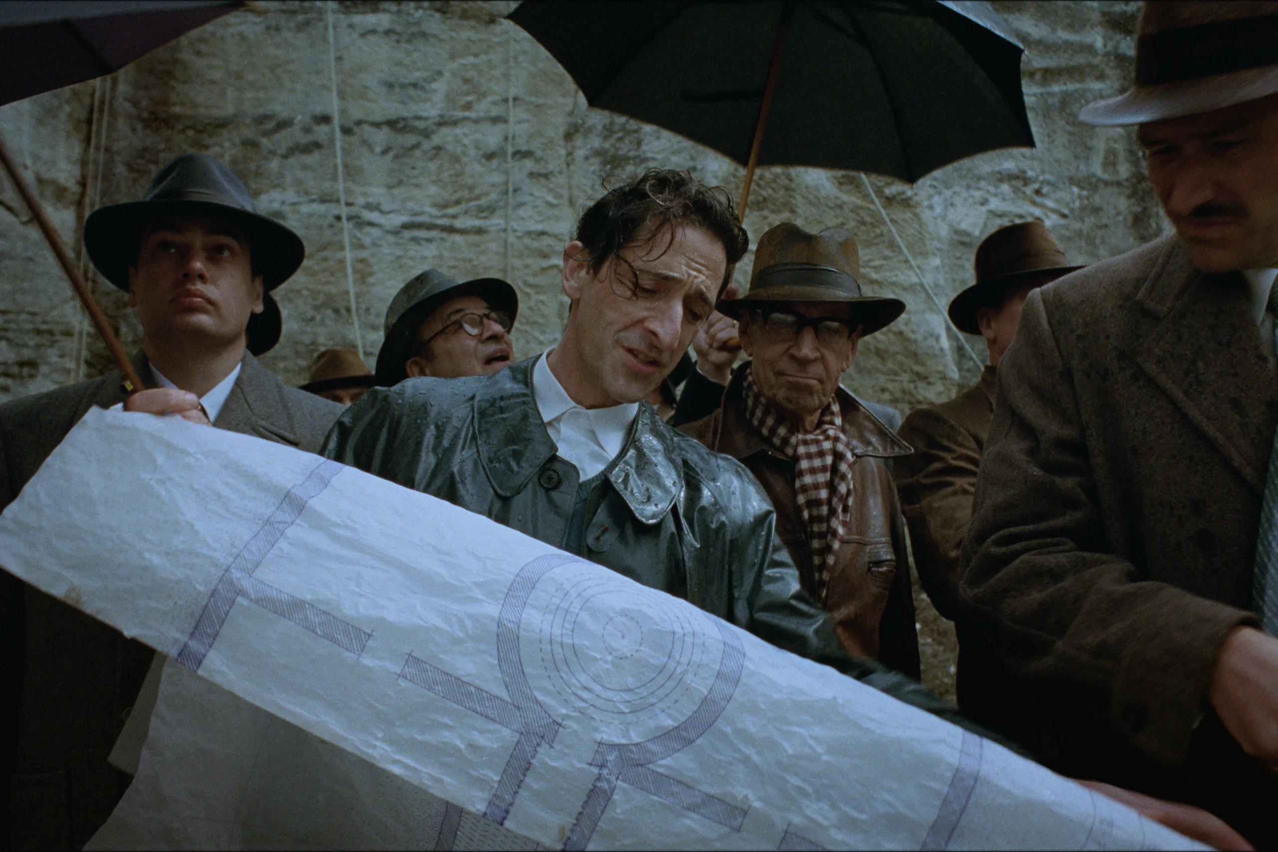 Adrien Brody looking over schematics while surrounded by men wearing coats and hats holding umbrellas in The Brutalist.