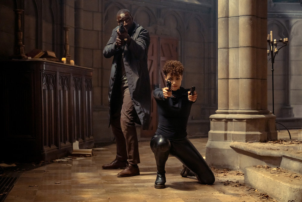 Omar Sy and Nathalie Emmanuel posing with a rifle and pistols in a church in The Killer.