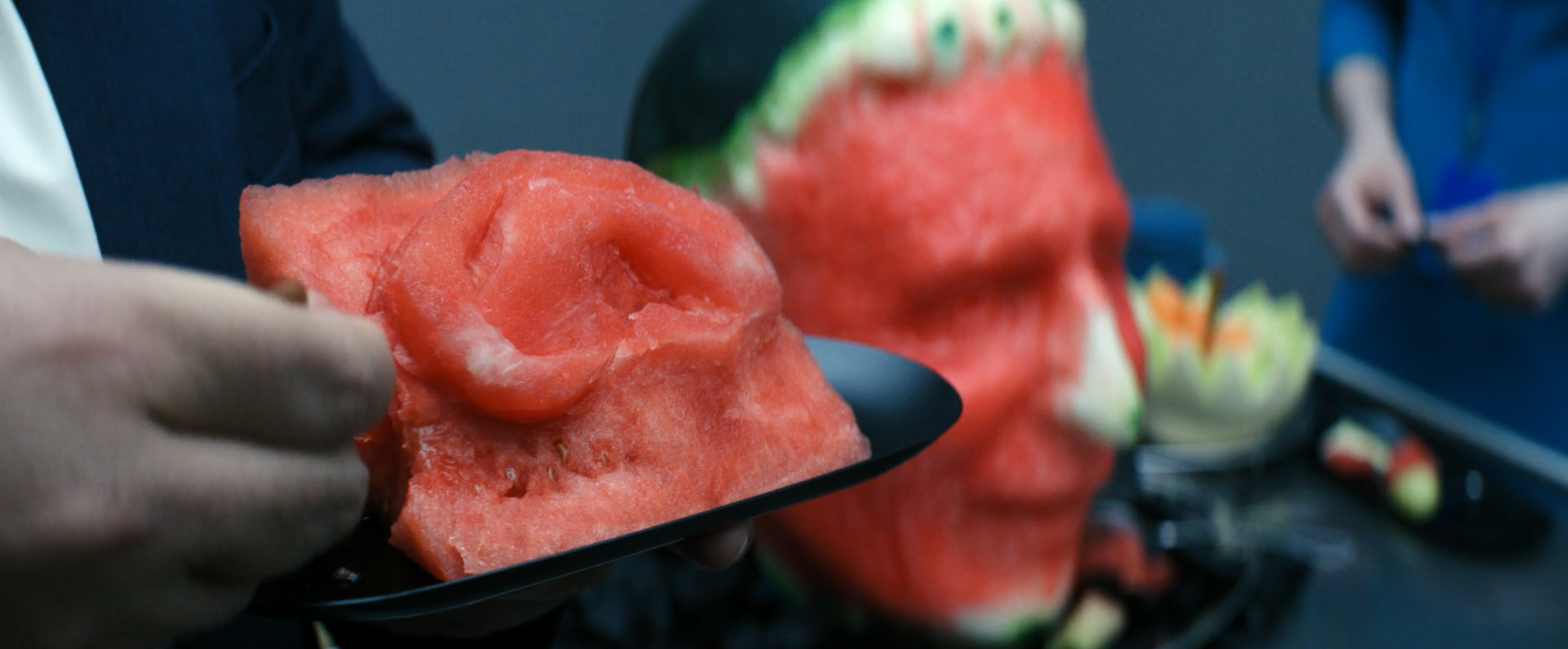 Dylan G eats a piece of watermelon from the Irving B fruit head sculpture in a still from Severance season 2, episode 5