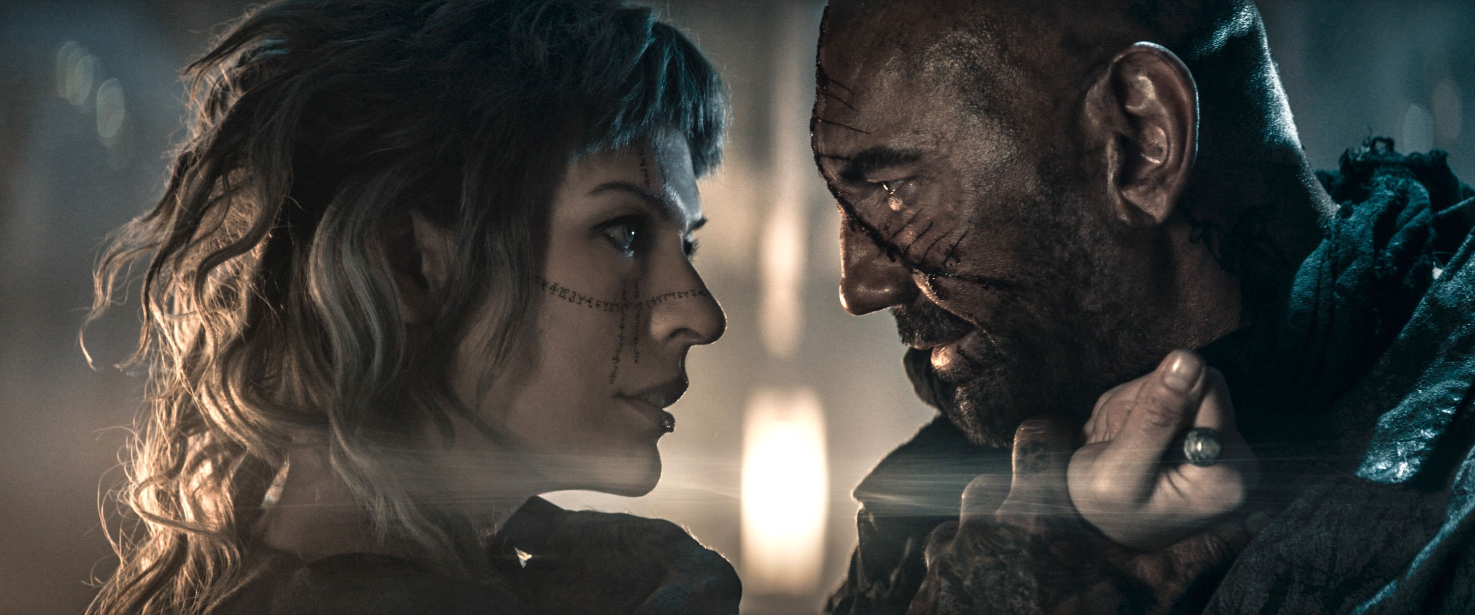 Gray Alys (Milla Jovovich) and Boyce the hunter (Dave Bautista), both grimy as hell and dramatically backlit, stand with their faces very close together, smiling just a little, in Paul W.S. Anderson’s In the Lost Lands