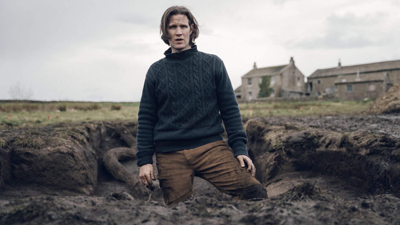 Matt Smith in a navy blue sweater standing in a large hole in the ground in Starve Acre.