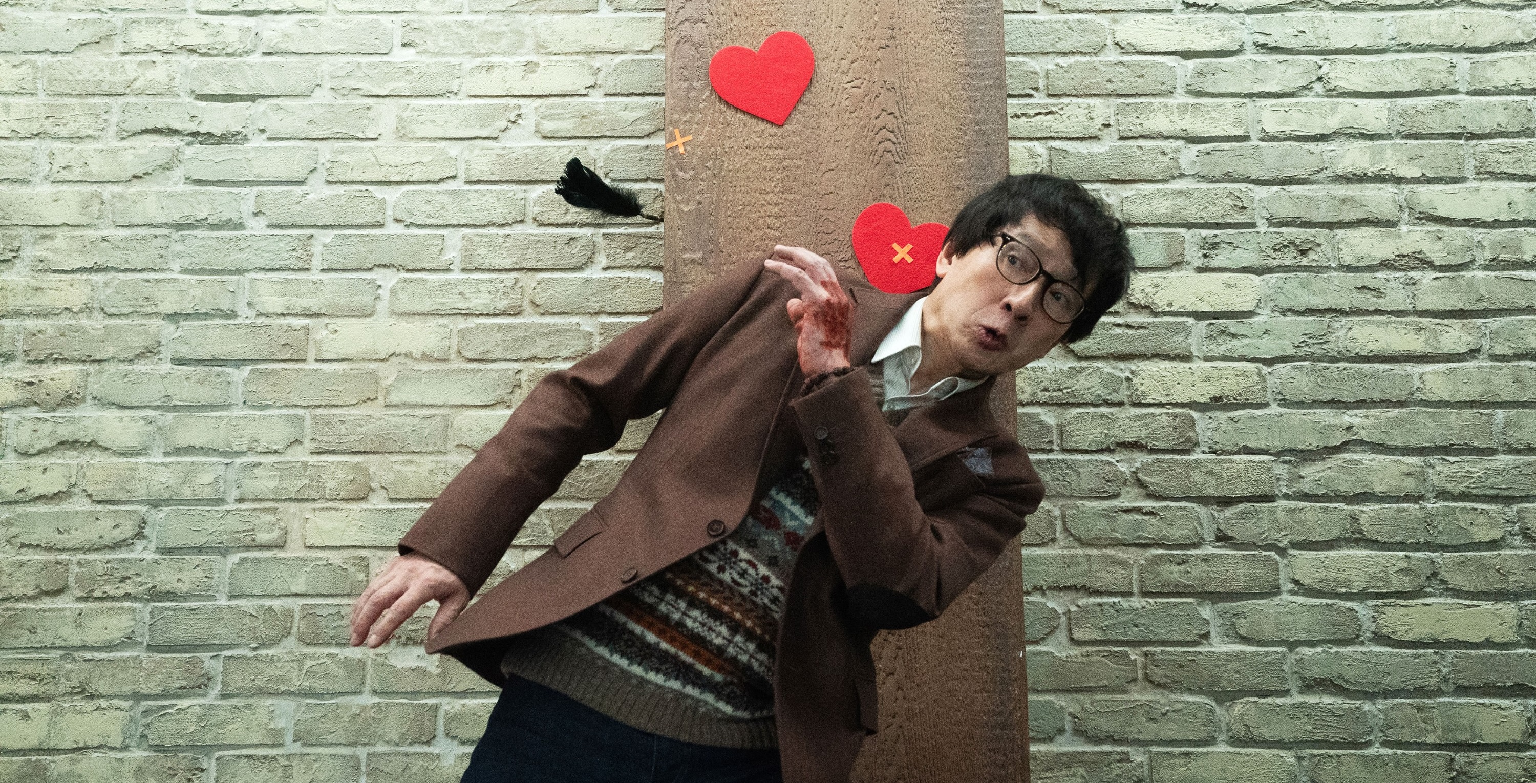 Marvin (Ke Huy Quan), a real-estate agent in brown jacket and striped sweater, stands in front og a brick wall dotted with paper heart cutouts and dodges a flying black feather with a comically exaggerated expression on his face in Love Hurts