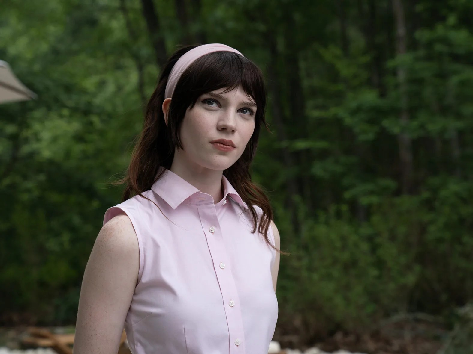 A brown haired woman in a short-sleeved, pink buttoned-up shirt and matching headband standing against the backdrop of a forest in Companion.