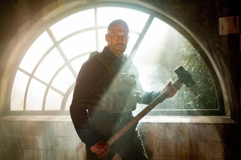 Jason Statham wearing an armored vest and holding a sledgehammer in front of a sunny half-circle window in A Working Man.