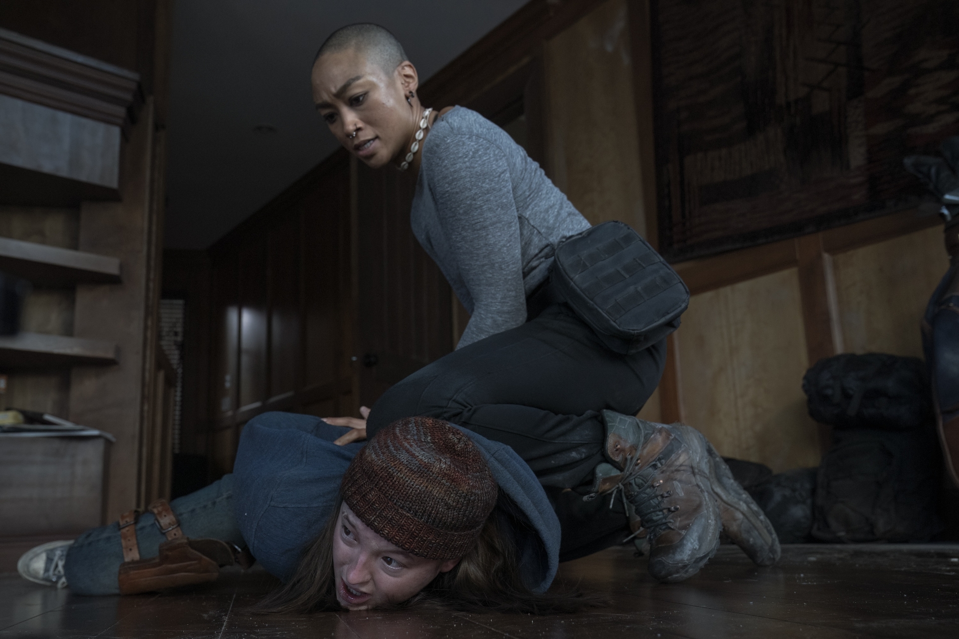 Bella Ramsey and Tati Gabrielle as Ellie and Nora. Nora is kneeling on Ellie who lies prone, and holds Ellie’s hands behind her back.