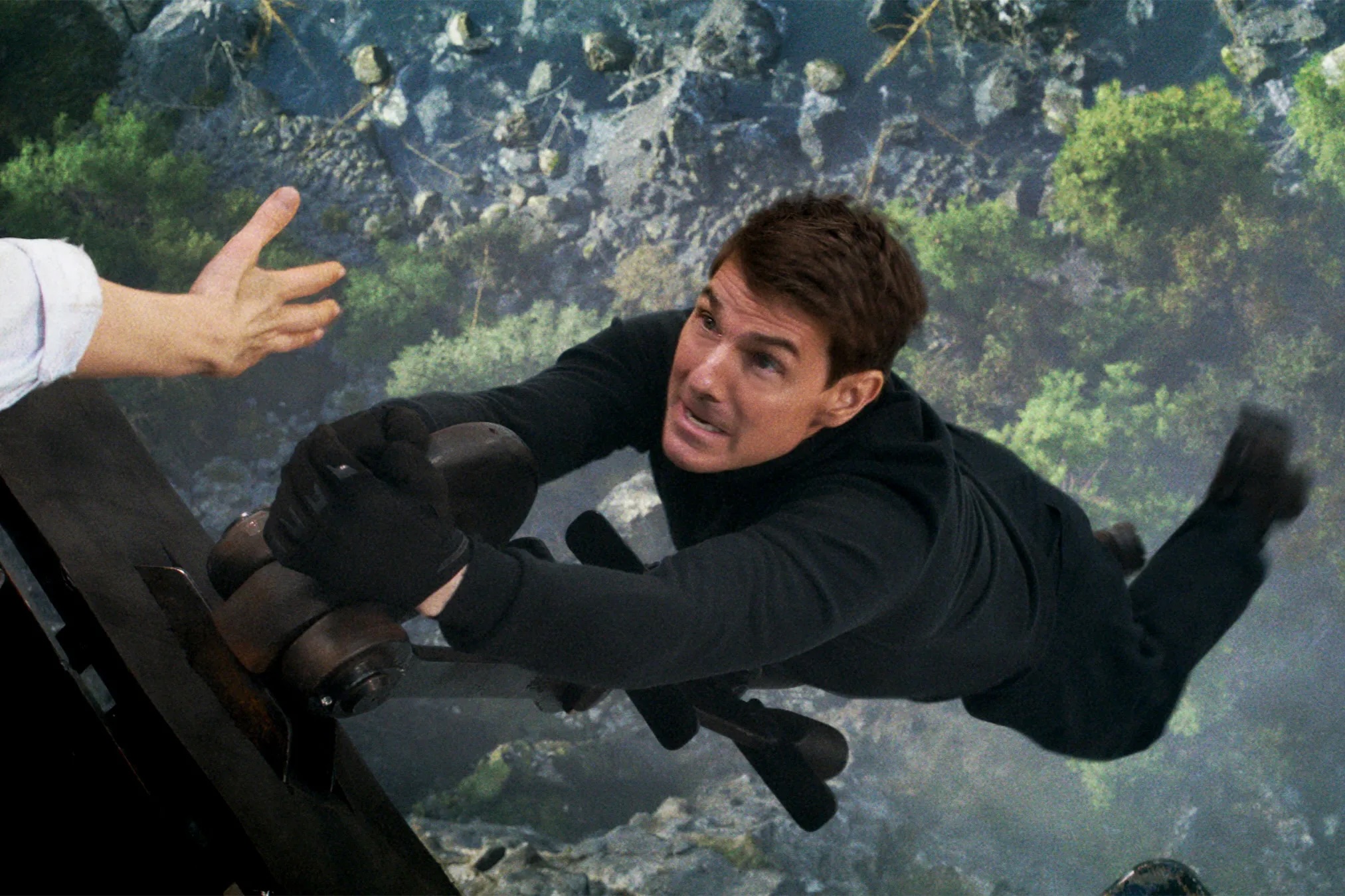 Ethan Hunt (Tom Cruise), in all black, hangs from the coupling of a train car over a river far below as an off-screen character reaches out to offer him a hand in Mission: Impossible — Dead Reckoning Part One