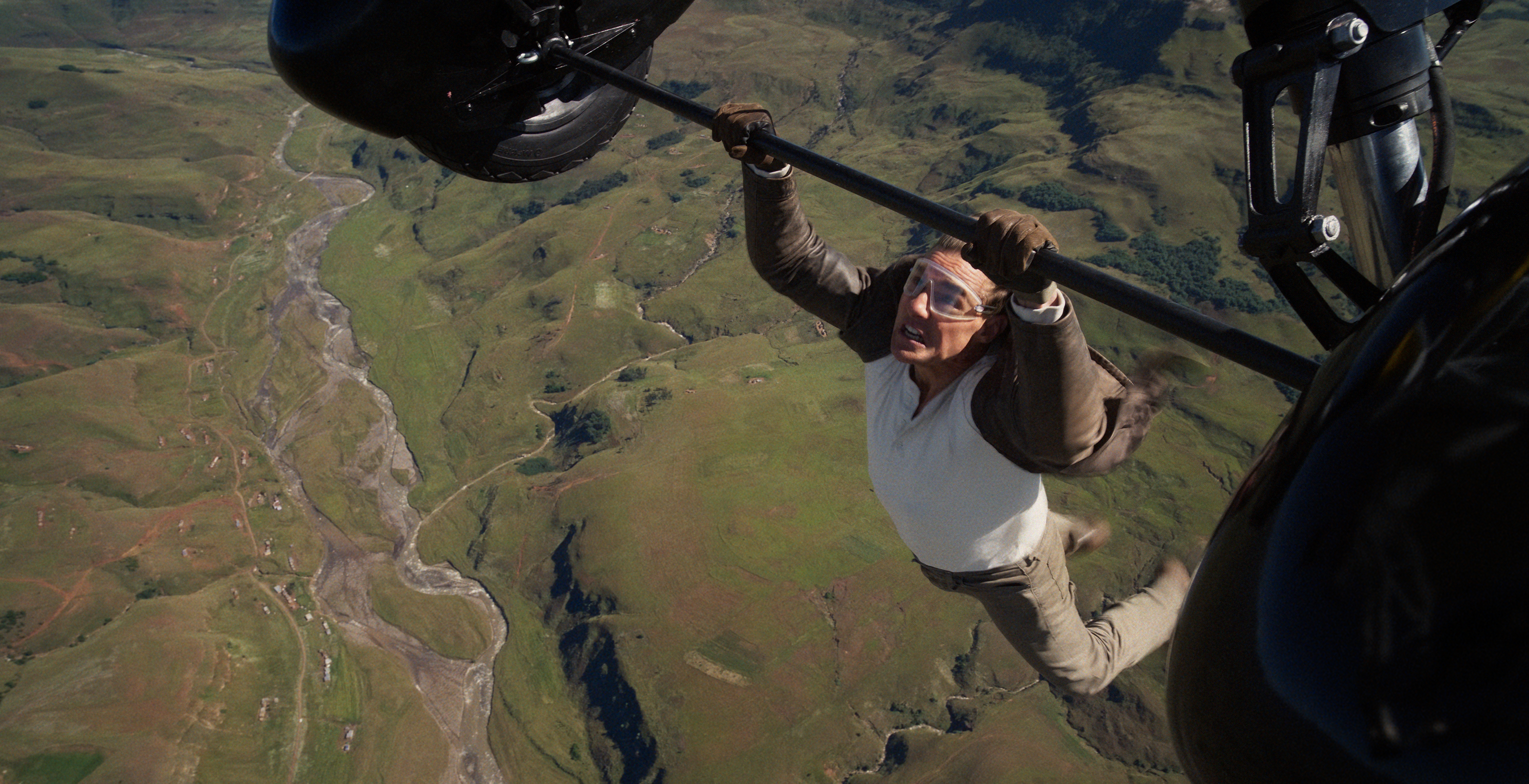 Ethan Hunt (Tom Cruise) hangs by his hands from the landing strut of a flying biplane, green fields in the distance far below, in Mission: Impossible – The Final Reckoning