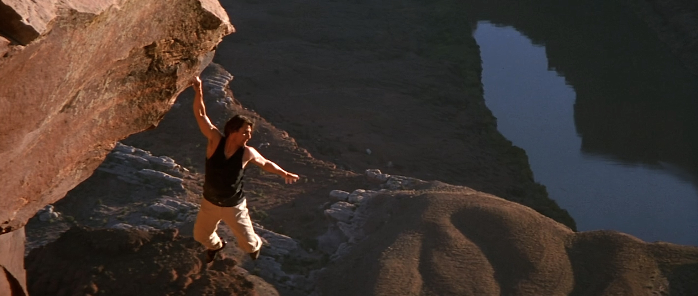 Ethan Hunt hangs from one hand from the edge of a raw red rock cliff, turning his body to look at the valley far below him, in Mission: Impossible II